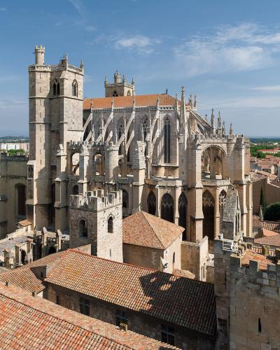 A high-angle photograph of a large, unfinished Gothic cathedral with a prominent square bell tower on the left, flying buttresses, and a terracotta-tiled roof, set within a dense urban environment of similar tiled rooftops under a blue sky.