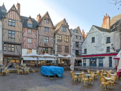 A cobblestone square lined with distinctive half-timbered medieval buildings and outdoor café seating under umbrellas, with a blue tarp-covered object in the center.