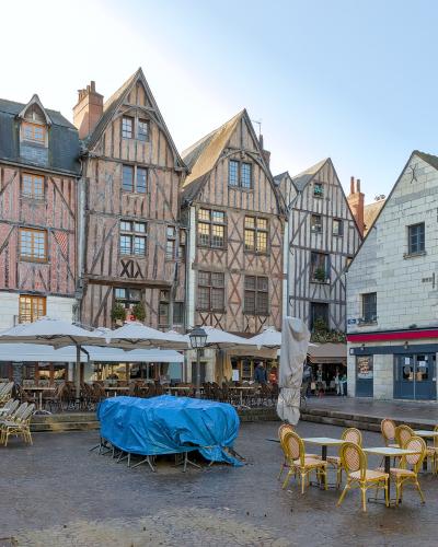 A cobblestone square lined with distinctive half-timbered medieval buildings and outdoor café seating under umbrellas, with a blue tarp-covered object in the center.