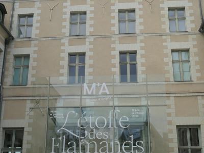 The image shows the modern glass entrance facade of a historic stone building, with signage reading "M'A Musées d'Angers" and exhibition titles including "L'étoffe des Flamands" and "Cultes et sanctuaires à Angers dans l'Antiquité," indicating it