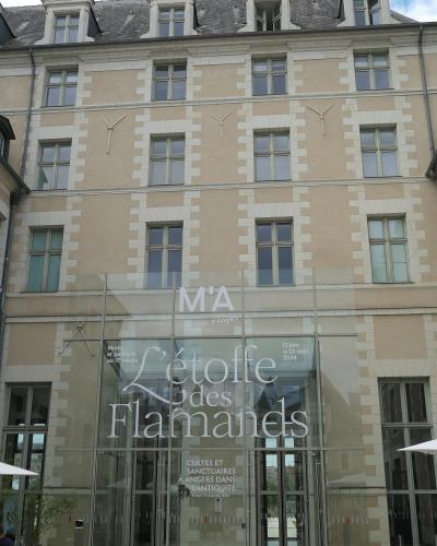 The image shows the modern glass entrance facade of a historic stone building, with signage reading "M'A Musées d'Angers" and exhibition titles including "L'étoffe des Flamands" and "Cultes et sanctuaires à Angers dans l'Antiquité," indicating it