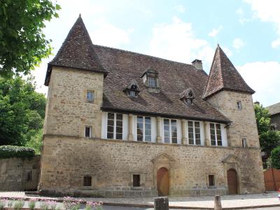 A large stone building with two conical towers, a tiled roof, and a row of tall windows on the upper floor, situated on a street with a car and trees nearby.