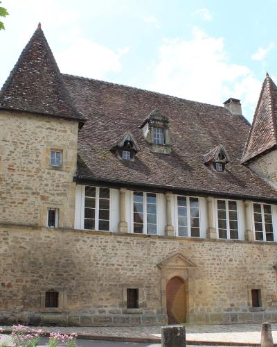 A large stone building with two conical towers, a tiled roof, and a row of tall windows on the upper floor, situated on a street with a car and trees nearby.