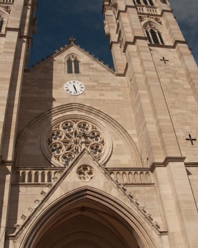 A low-angle photograph of a large stone cathedral facade featuring two prominent square towers, a central rose window, a clock, and a Gothic-style arched entrance.