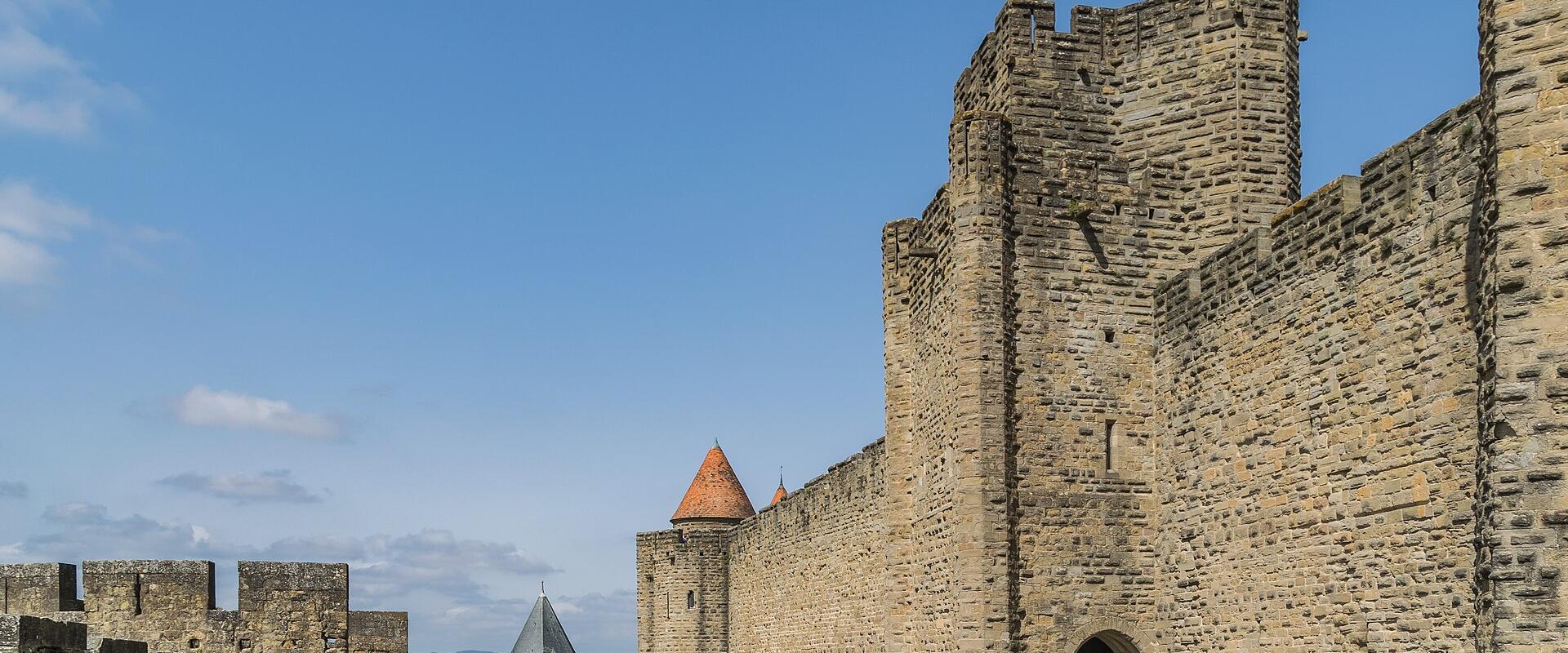The image shows the stone ramparts and towers of a medieval fortified city under a blue sky, with tourists walking along the walkways and near an arched gateway; a conical-roofed tower and distant landscape are visible in the background.