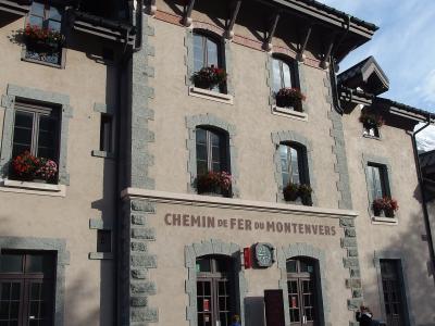 A beige stone-and-plaster building with arched windows, flower boxes, and the sign "CHEMIN DE FER DU MONTENVERS" above the entrance doors, with people standing outside on a paved street under a partly cloudy sky.