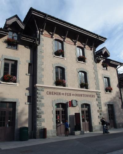 A beige stone-and-plaster building with arched windows, flower boxes, and the sign "CHEMIN DE FER DU MONTENVERS" above the entrance doors, with people standing outside on a paved street under a partly cloudy sky.