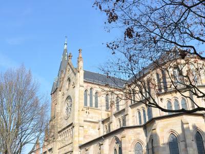 A low-angle exterior shot of a large Neo-Gothic stone church with a prominent rose window, pointed arches, and a steep slate roof, viewed from a street corner with bare trees and a pedestrian crossing in the foreground.
