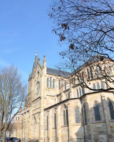 A low-angle exterior shot of a large Neo-Gothic stone church with a prominent rose window, pointed arches, and a steep slate roof, viewed from a street corner with bare trees and a pedestrian crossing in the foreground.