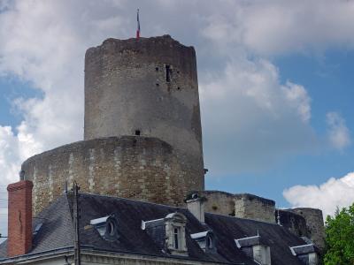 A tall, cylindrical medieval stone tower with a French flag on top, rising above a slate-roofed building with dormer windows and a brick chimney, under a partly cloudy sky.