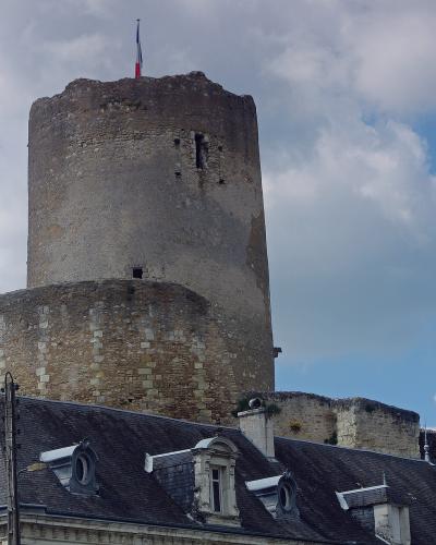 A tall, cylindrical medieval stone tower with a French flag on top, rising above a slate-roofed building with dormer windows and a brick chimney, under a partly cloudy sky.