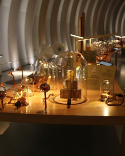 An indoor museum exhibit featuring copper-colored wine-related artifacts and glass domes on a wooden table, set against a distinctive curved, ribbed white wall with recessed lighting.