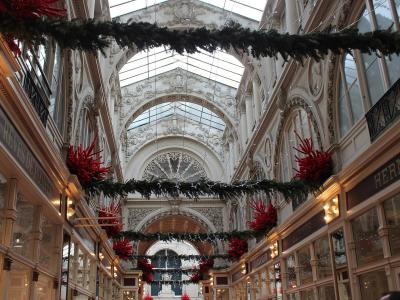 The image shows the interior of a grand, ornate 19th-century shopping arcade with a glass roof, elaborate plasterwork, and Christmas decorations including garlands and red coral-like ornaments; visible signage includes “RÊVES DE POMMES” and “HERMÈ