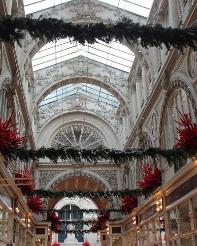The image shows the interior of a grand, ornate 19th-century shopping arcade with a glass roof, elaborate plasterwork, and Christmas decorations including garlands and red coral-like ornaments; visible signage includes “RÊVES DE POMMES” and “HERMÈ