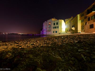 A low-angle night photograph of a pebble beach with illuminated multi-story buildings along the waterfront, showing a starry sky and distant city lights across the water.