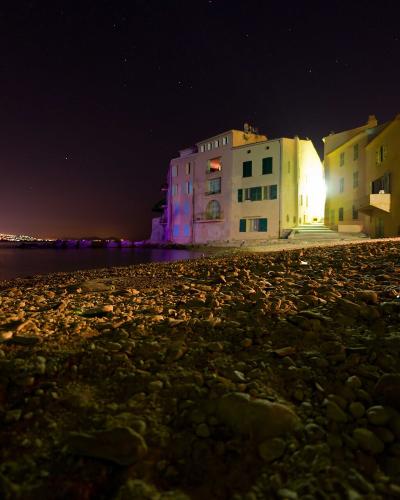 A low-angle night photograph of a pebble beach with illuminated multi-story buildings along the waterfront, showing a starry sky and distant city lights across the water.