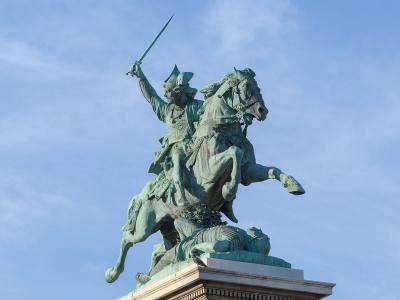 A bronze equestrian statue of a warrior on horseback, raising a sword, with a fallen figure beneath the horse’s hooves, set against a blue sky; the statue stands on a stone pedestal with classical architectural details.