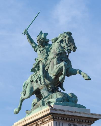 A bronze equestrian statue of a warrior on horseback, raising a sword, with a fallen figure beneath the horse’s hooves, set against a blue sky; the statue stands on a stone pedestal with classical architectural details.