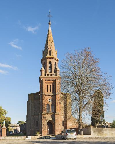 A red brick church with a tall, pointed spire and a bell tower, set against a blue sky with a large tree to the right and a silver car parked in front.