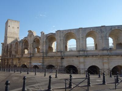The image shows the exterior of a large, ancient Roman amphitheater with two tiers of arches, a prominent square tower on the left, and modern black bollards in the foreground under a clear blue sky.