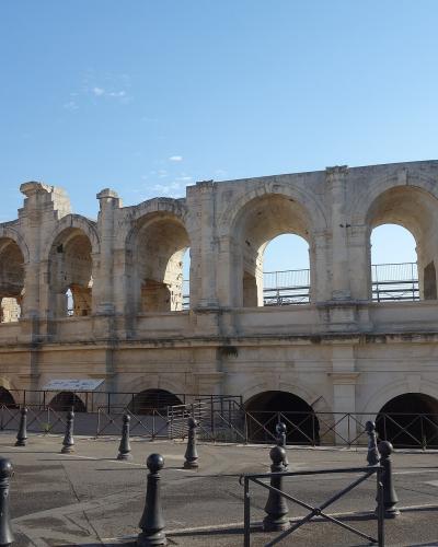 The image shows the exterior of a large, ancient Roman amphitheater with two tiers of arches, a prominent square tower on the left, and modern black bollards in the foreground under a clear blue sky.