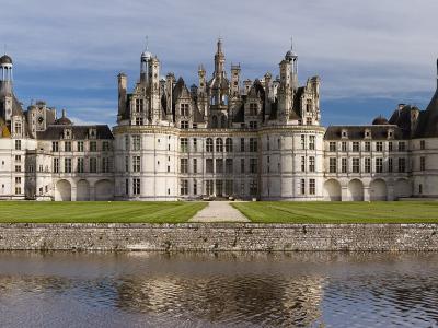 1. A wide-angle photograph of a large, ornate Renaissance château with distinctive French medieval towers and chimneys, situated on a grassy bank above a moat or river under a partly cloudy sky.