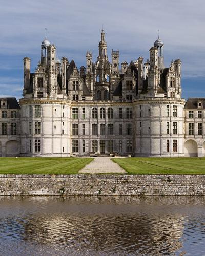 1. A wide-angle photograph of a large, ornate Renaissance château with distinctive French medieval towers and chimneys, situated on a grassy bank above a moat or river under a partly cloudy sky.