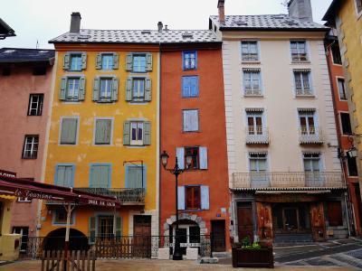 A street-level view of colorful, multi-story buildings with shuttered windows and balconies in a European-style town square, featuring a red awning labeled "le Tourisme" and a black lamppost.
