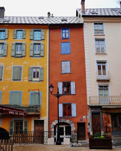 A street-level view of colorful, multi-story buildings with shuttered windows and balconies in a European-style town square, featuring a red awning labeled "le Tourisme" and a black lamppost.