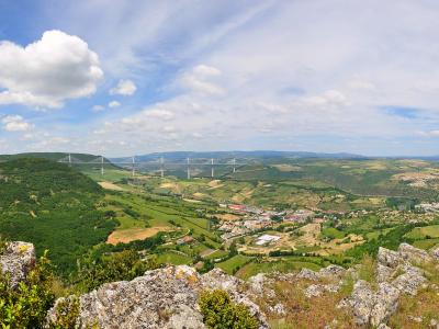 A wide landscape photograph showing a long cable-stayed bridge with multiple tall, slender pylons spanning a deep green valley, viewed from a rocky foreground under a partly cloudy sky.