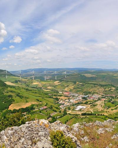 A wide landscape photograph showing a long cable-stayed bridge with multiple tall, slender pylons spanning a deep green valley, viewed from a rocky foreground under a partly cloudy sky.