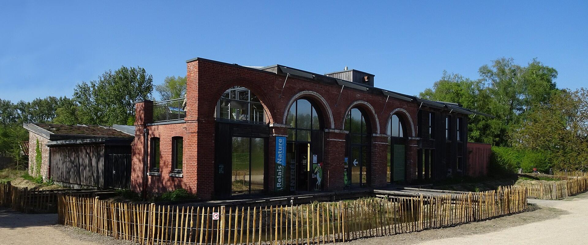 A red brick building with large arched windows and a blue vertical sign reading "Relais Nature" and "ENTREE", surrounded by a wooden fence and trees under a clear blue sky.
