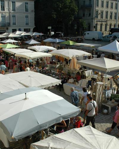 An outdoor market with numerous white umbrellas and stalls set up on a paved square, with classical European-style buildings in the background and people browsing.