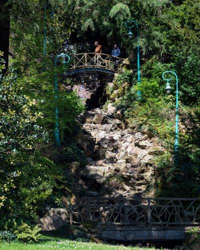 Two people stand on an arched bridge overlooking a rocky grotto with turquoise lampposts in a lush green park.