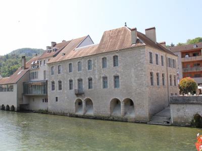 A large, multi-story stone building with arched openings at its base along a riverbank, featuring a modern glass extension on the left and a tiled roof; no visible signage or text is present in the image.