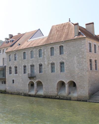 A large, multi-story stone building with arched openings at its base along a riverbank, featuring a modern glass extension on the left and a tiled roof; no visible signage or text is present in the image.