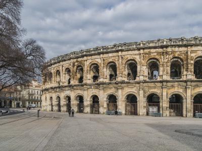 A wide-angle view of the Arènes de Nîmes, showing its two-tiered Roman amphitheater façade with arched openings, set against a cloudy sky, with a paved plaza in the foreground and bare trees to the left.
