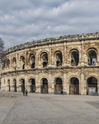 A wide-angle view of the Arènes de Nîmes, showing its two-tiered Roman amphitheater façade with arched openings, set against a cloudy sky, with a paved plaza in the foreground and bare trees to the left.