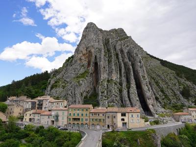 A large, distinctive limestone rock formation (the Rocher de la Baume) towering over the village of Saignon, with buildings and a bridge in the foreground.