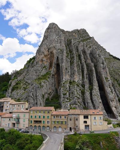 A large, distinctive limestone rock formation (the Rocher de la Baume) towering over the village of Saignon, with buildings and a bridge in the foreground.