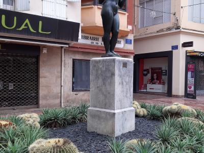 A bronze statue of a nude female figure standing on a stone pedestal, surrounded by cacti and succulents in a paved urban plaza, with storefronts including "LIBRAIRIE de la Loge" and "NOCIBÉ" visible in the background.