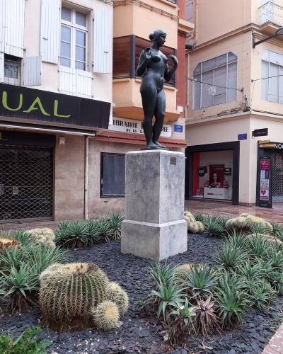 A bronze statue of a nude female figure standing on a stone pedestal, surrounded by cacti and succulents in a paved urban plaza, with storefronts including "LIBRAIRIE de la Loge" and "NOCIBÉ" visible in the background.