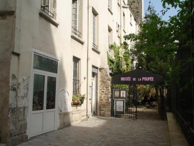 A narrow cobblestone alleyway in a historic European town, featuring a black canopy tent with the text "MUSÉE DE LA POUPÉE" in pink letters, situated next to a white-walled building with stone foundations and a window displaying dolls.