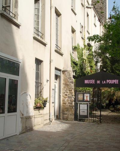 A narrow cobblestone alleyway in a historic European town, featuring a black canopy tent with the text "MUSÉE DE LA POUPÉE" in pink letters, situated next to a white-walled building with stone foundations and a window displaying dolls.