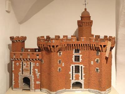 A detailed architectural model of a red-brick fortified gatehouse with crenellations, a central tower topped by a dome and weather vane, white stone accents around windows and arches, and a small striped flag hanging on the right side, displayed i