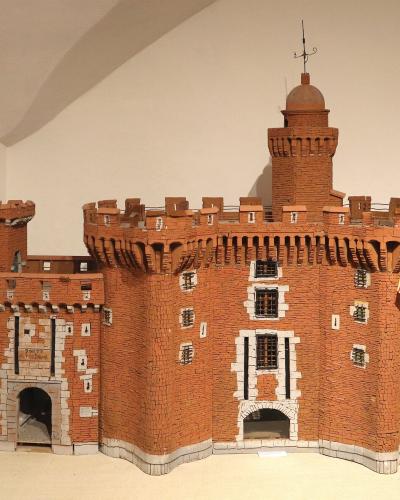 A detailed architectural model of a red-brick fortified gatehouse with crenellations, a central tower topped by a dome and weather vane, white stone accents around windows and arches, and a small striped flag hanging on the right side, displayed i