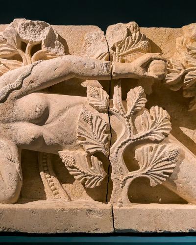 A stone relief sculpture depicting a reclining nude female figure reaching toward a tree laden with fruit and leaves, displayed against a dark background on a blue surface.