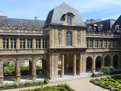 A black-and-white photograph of a formal courtyard garden with ornate gravel patterns, flanked by Renaissance-style buildings featuring arched colonnades and classical facades, with a watermark in the lower-left corner reading "PALIER" and "PARIS"