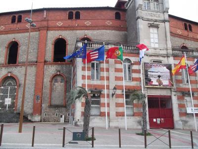 The image shows the exterior facade of a large brick and stone arena building with arched entryways, multiple flags (including French, Portuguese, Spanish, and EU flags), and a prominent sign reading "BÉZIERS LES ARÈNES" above a red gate; the word