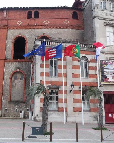 The image shows the exterior facade of a large brick and stone arena building with arched entryways, multiple flags (including French, Portuguese, Spanish, and EU flags), and a prominent sign reading "BÉZIERS LES ARÈNES" above a red gate; the word